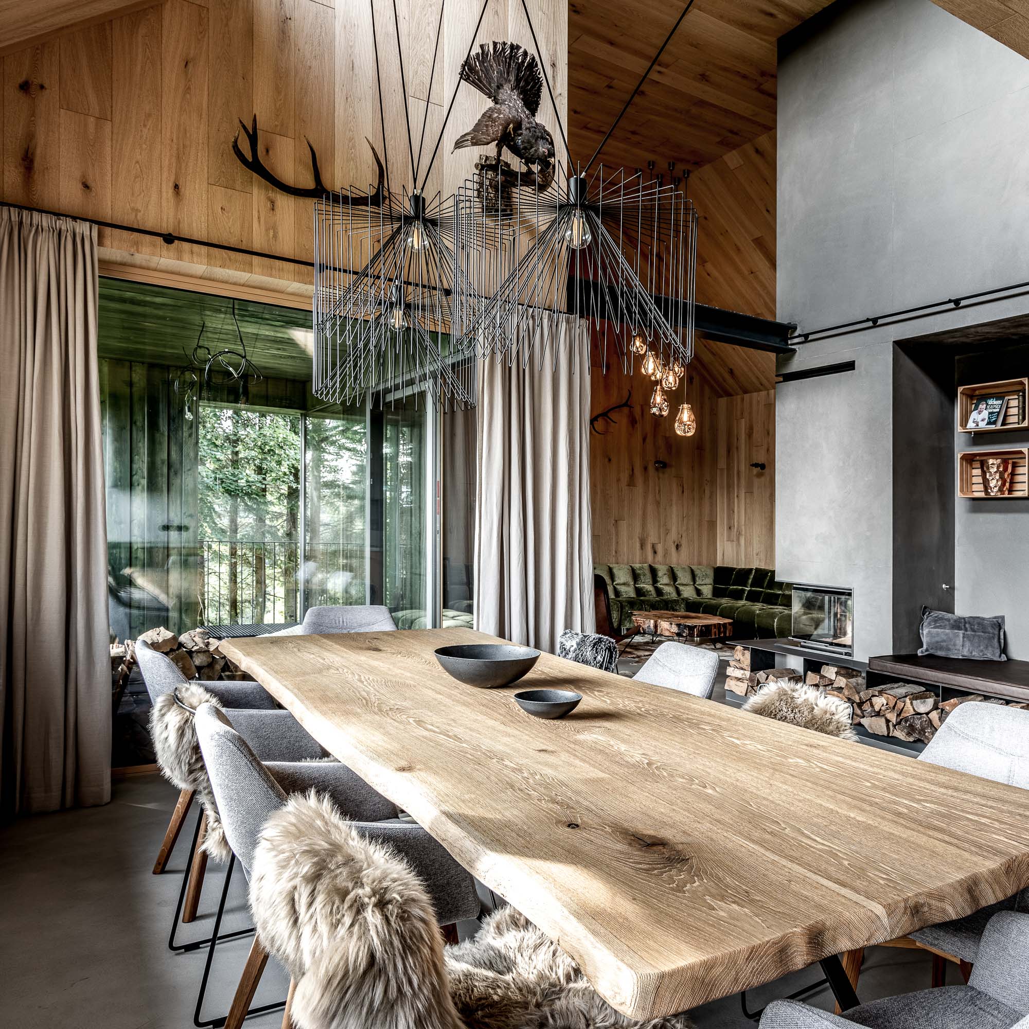 View from the head of the dining table across the open space of MARIÁNSKÁ chalet – starburst and amber pendant chandeliers, oak table with grey and fur chairs, living room and fireplace in background