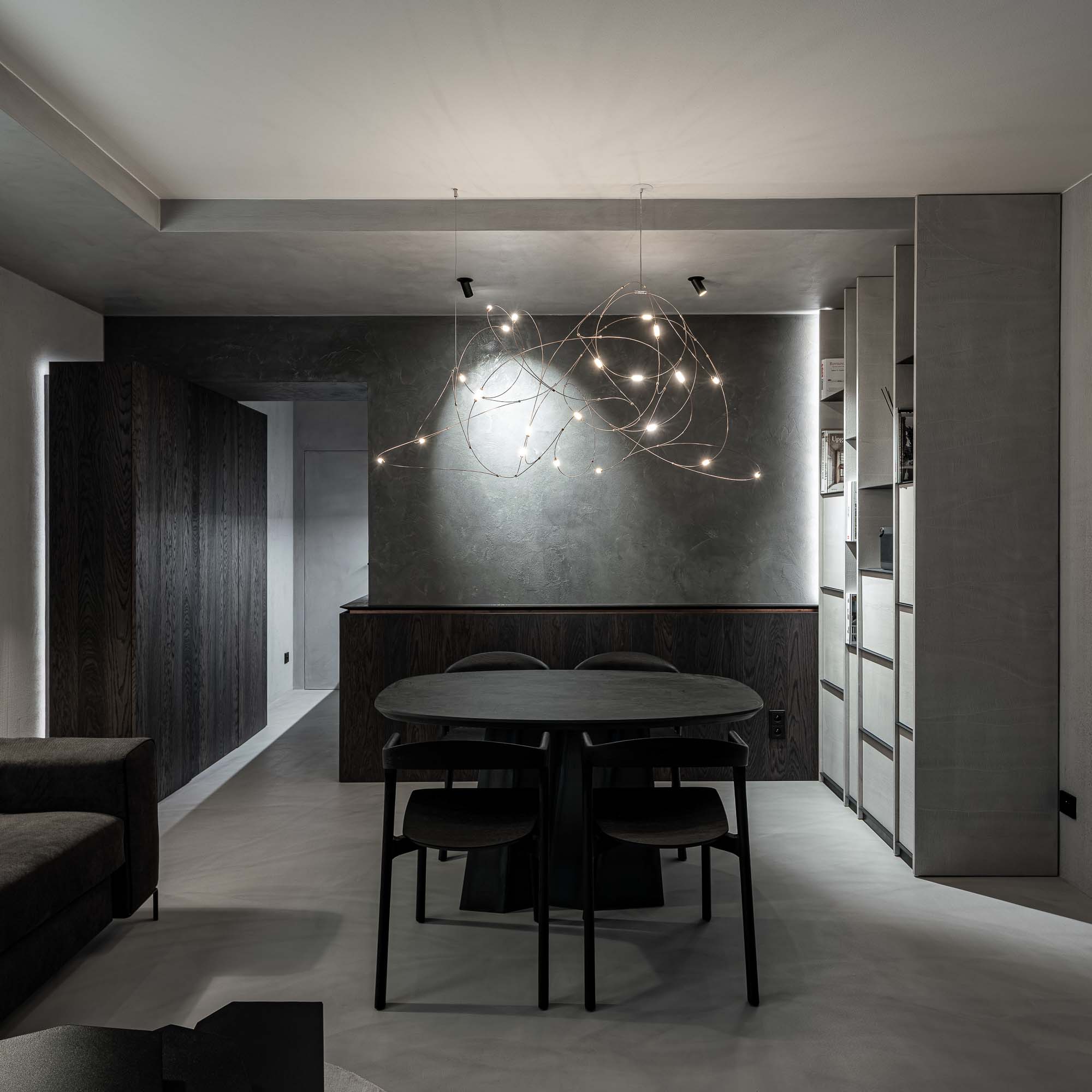 Centrální prostor lounge Selection Properties — černý oválný stůl Ethnicraft Bok se čtyřmi křesly, lustr Moooi Flock of Light, tmavá stěrkovaná stěna, vestavná knihovna vpravo, kuchyňka v pozadí vlevo